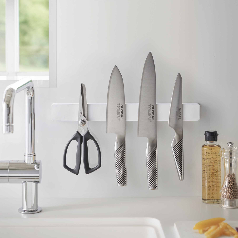 White Magnetic Knife Rack from Yamazaki Home holding a set of kitchen knives and scissors in a modern kitchen. Chrome sink to the left and yellow soap to the right of the rack.