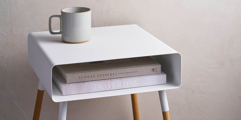 White steel storage table side table holding two coffee table books on the inner shelf and a mug on top. 