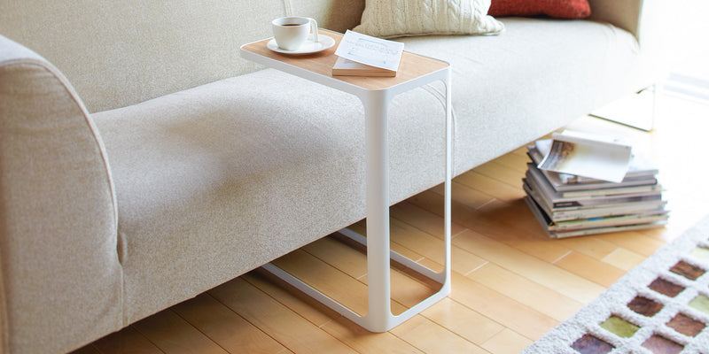 Yamazaki Home C Side Table in white and ash, tucked into a beige couch in a modern living room holding a cup of coffee and a book