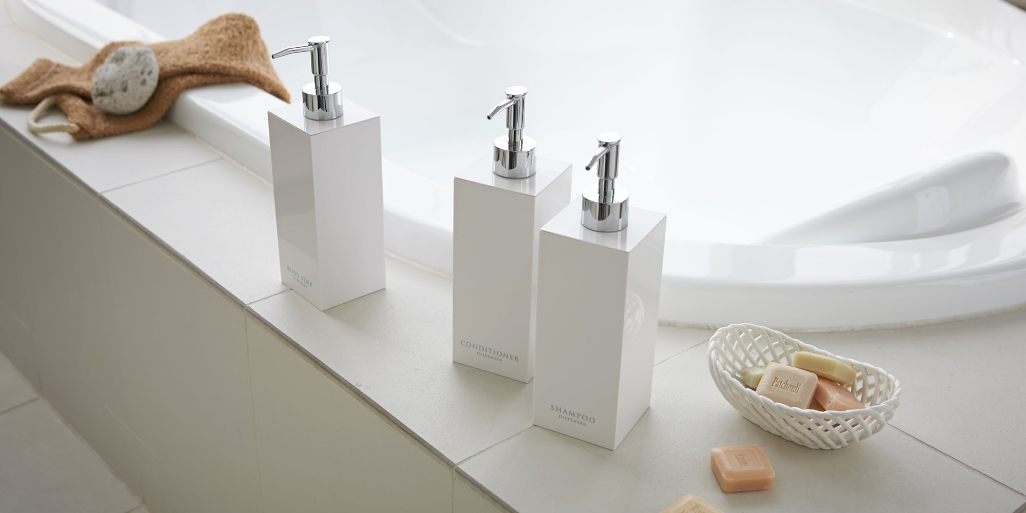 Yamazaki Home square soap dispensers on the side of a modern bathtub. Also featuring a bowl of patchouli soap and stone exfoliator.