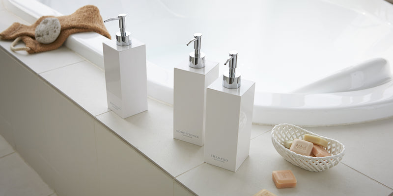 Yamazaki Home square soap dispensers on the side of a modern bathtub. Also featuring a bowl of patchouli soap and stone exfoliator.