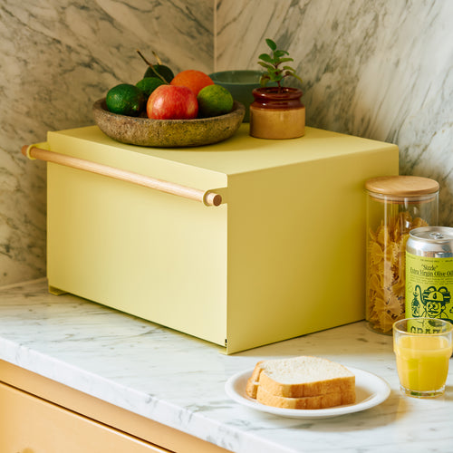 A pear yellow kitchen bread box with a wooden handle sits on a marble countertop, topped with a bowl of fresh fruit and a small potted plant.
