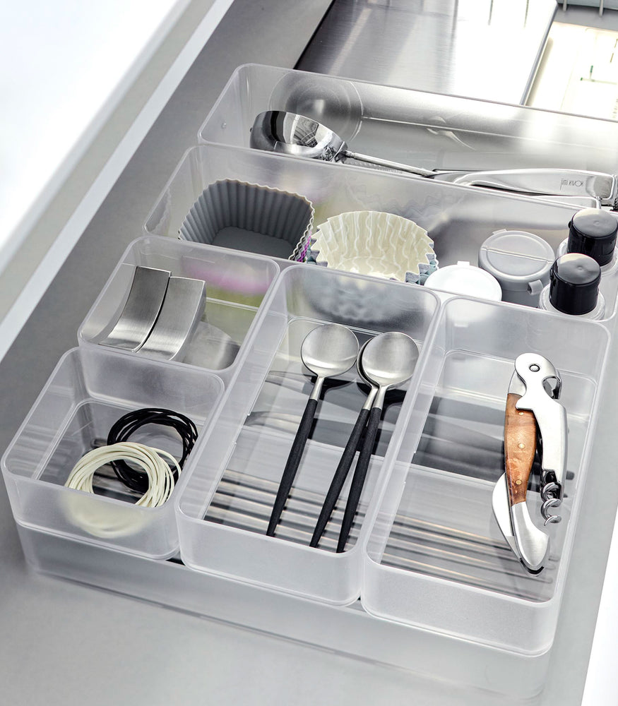 View 7 - Close-up image of four different sizes of stackable clear organizers—small, medium, large, and deep—neatly placed in a bathroom drawer. Made from transparent material, they accommodate items like brushes and sanitary products, showcasing their clarity and stackability.