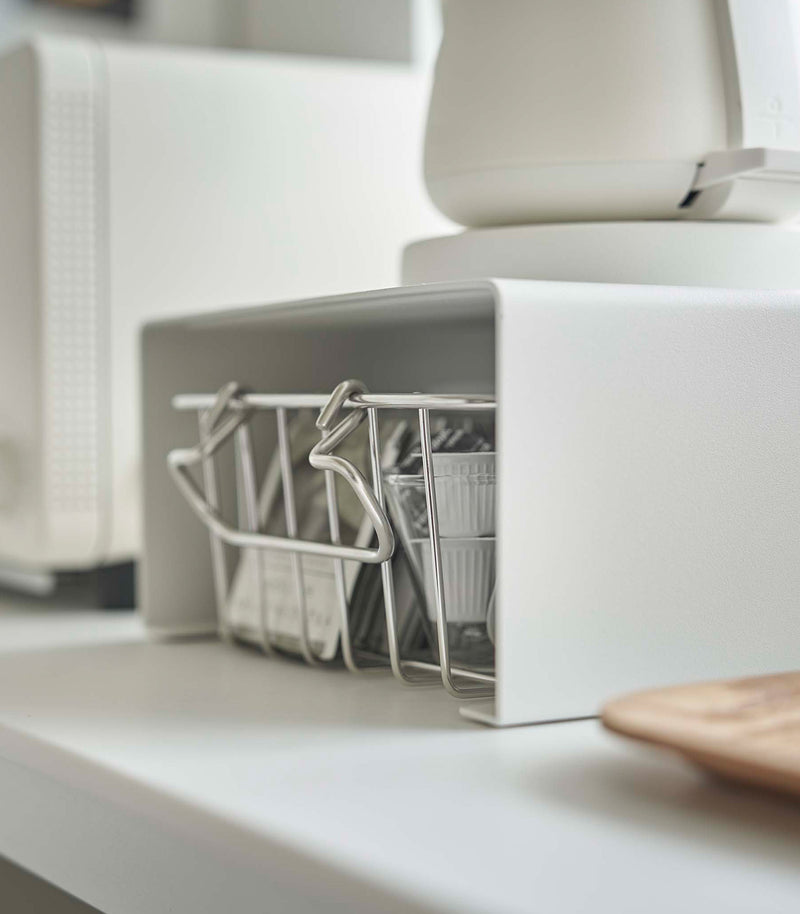 Close up of white Yamazaki Home Stackable Countertop Shelf with a basket of tea underneath and a kettle above