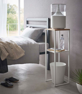 Pedestal Stand by Yamazaki Home in white in a bedroom holding a humidifier and books, and a waste bin underneath. view 3
