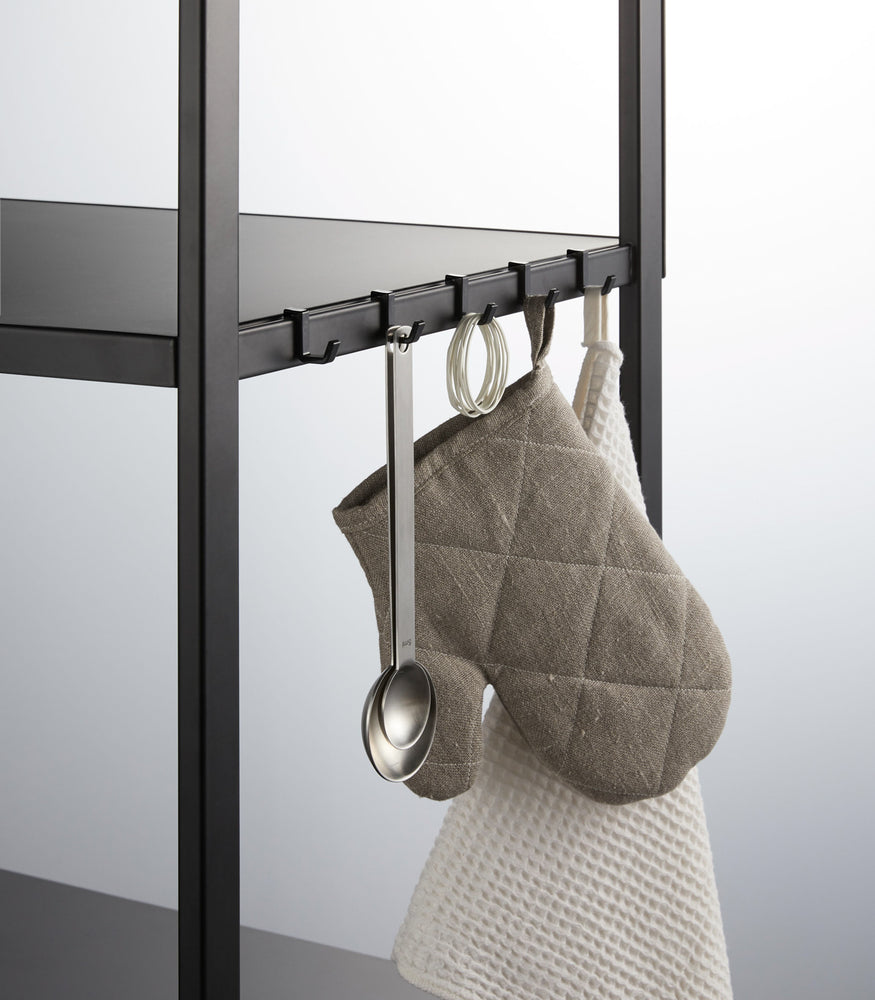 View 18 - Close of black Storage Rack hooks holding meauring tools and oven mits on white background by Yamazaki Home.