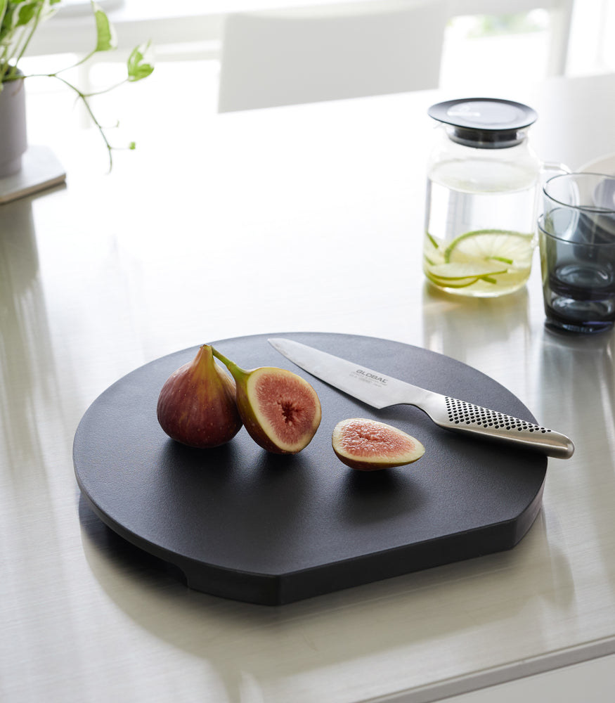 View 16 - A black Yamazaki Round Magnetic Cutting Board with a fig and knife on it on a countertop.