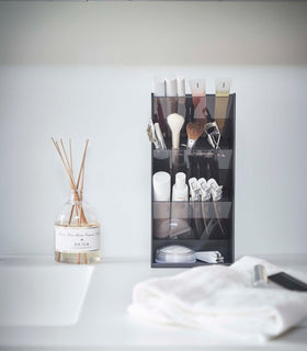 A front view of a black rectangular resin cosmetics organizer on a white bathroom counter. It has three deep adjustable black transparent trays that sit diagonally with matching dividers placed in the middle of each tray. view 29