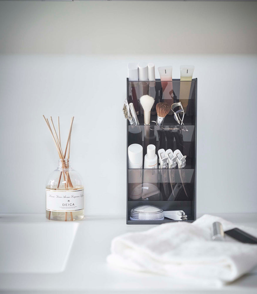 View 30 - A front view of a black rectangular resin cosmetics organizer on a white bathroom counter. It has three deep adjustable black transparent trays that sit diagonally with matching dividers placed in the middle of each tray.