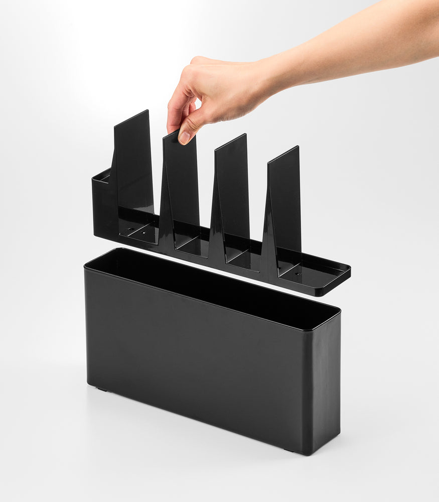 View 14 - Black Utensil & Thin Cutting Board Holder by Yamazaki Home with a hand placing a cutting board into one of its slots.