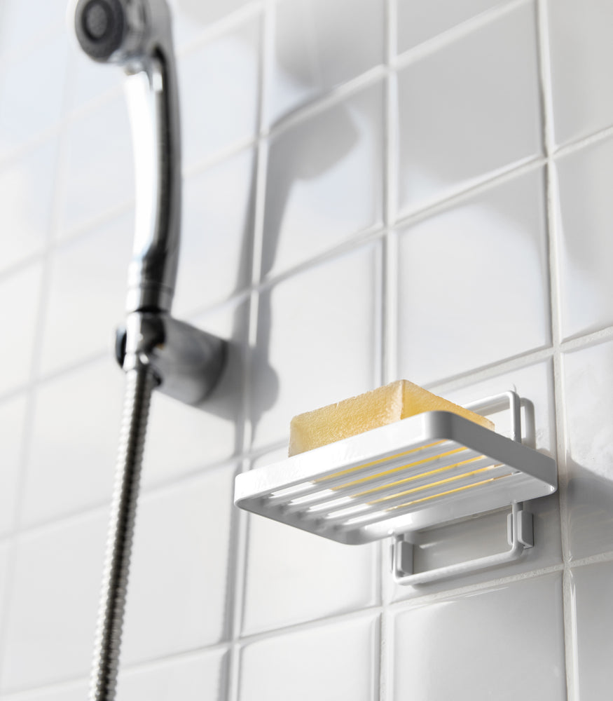 View 5 - Yamazaki Home's white Traceless Adhesive Soap Tray in a shower, holding a bar of soap next to a silver shower hose on tiled wall.