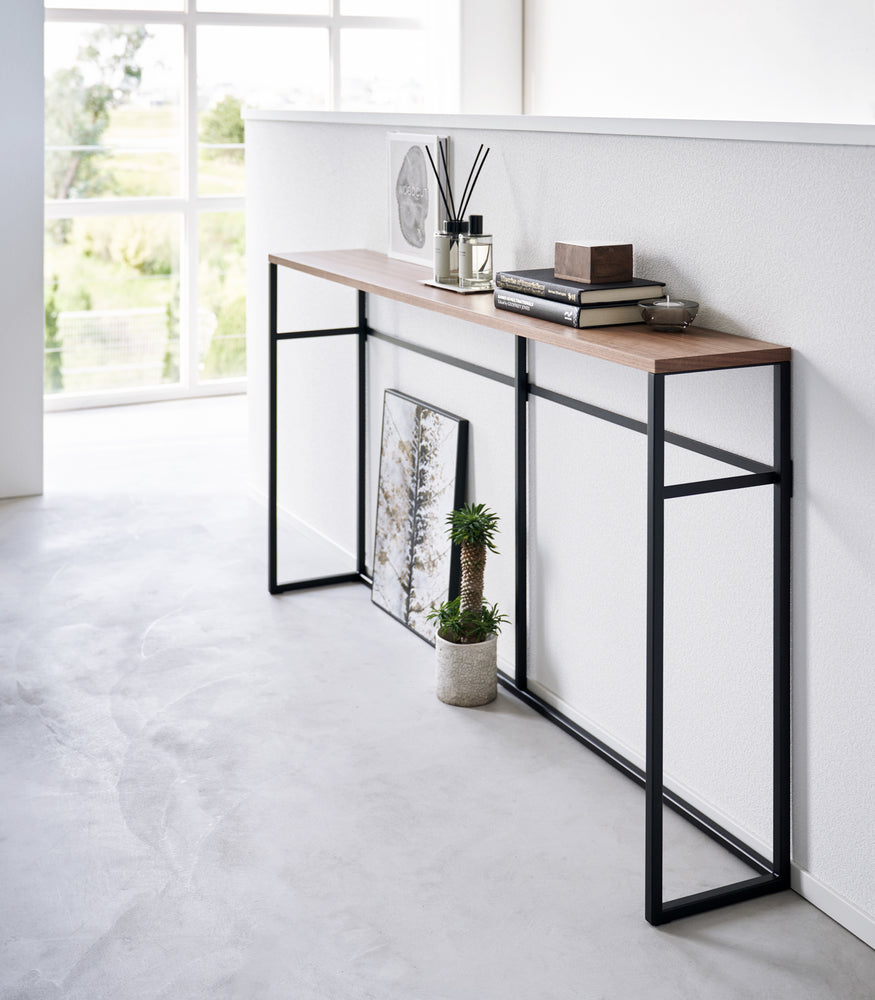 View 11 - Diagonal view of the Long Console Table by Yamazaki Home in black placed by a wall with decorative items.