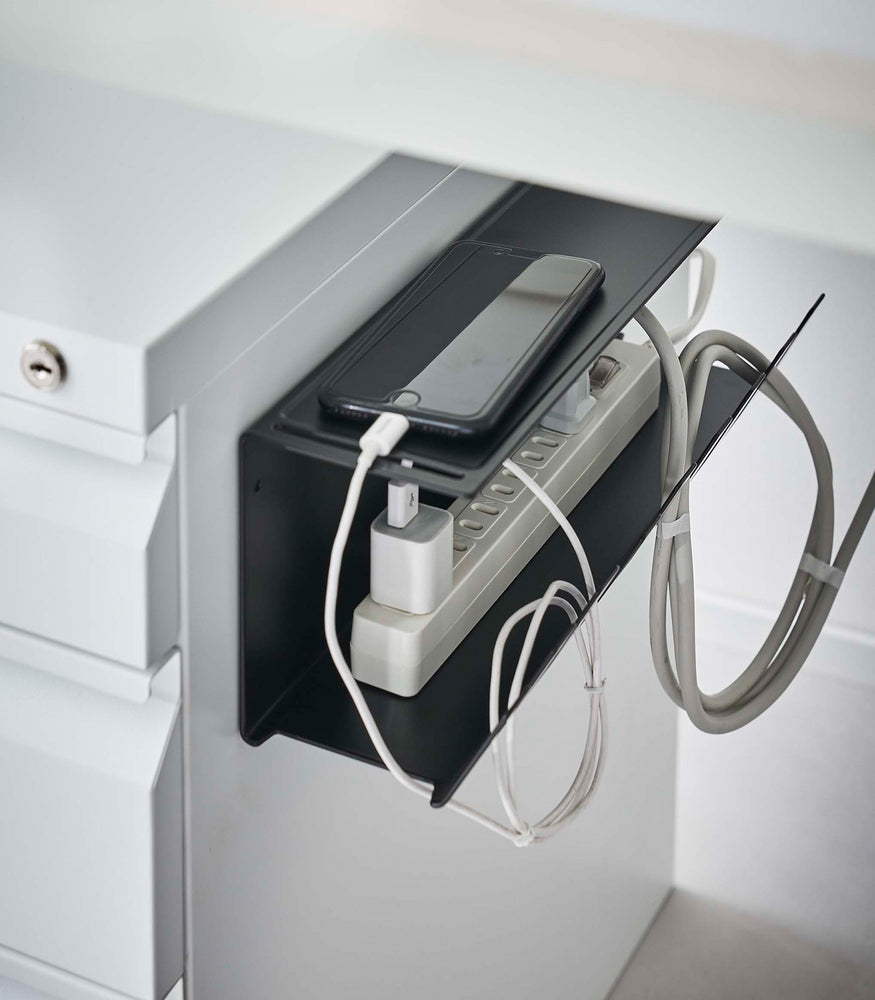 View 16 - Close-up of Magnetic Under-Desk Cable Organizer in white by Yamazaki Home mounted on the side of a file cabinet holding a power strip and phone.