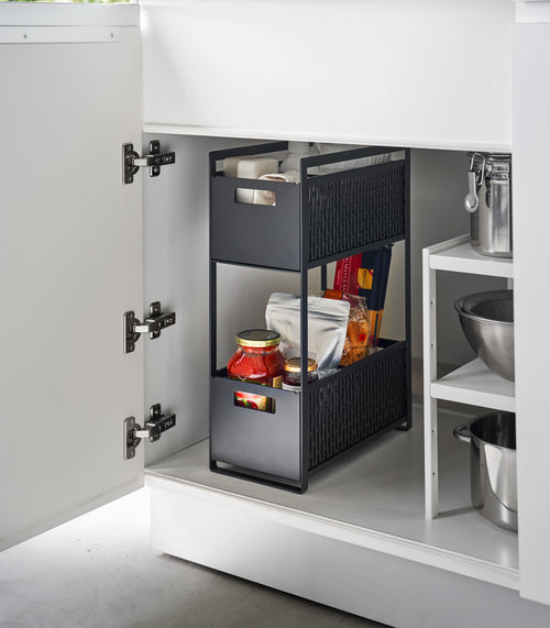 A two-tier black storage bin is shown in a lower cabinet. The upper level holds cleaning supplies, the bottom level holds pantry items.