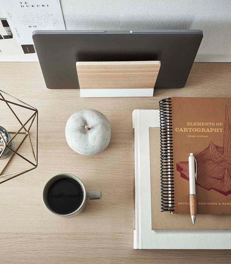 A wooden laptop stand with a minimalist white metal base rests behind an assortment of desk items, captured from an overhead perspective.