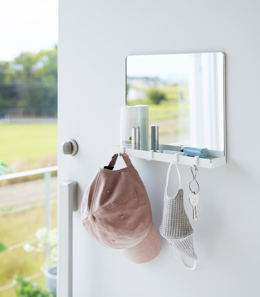 View 6 - A white Yamazaki Magnetic Mirror With Storage Rack attached to a door with a hanging hat, mask, and key with a view outside.