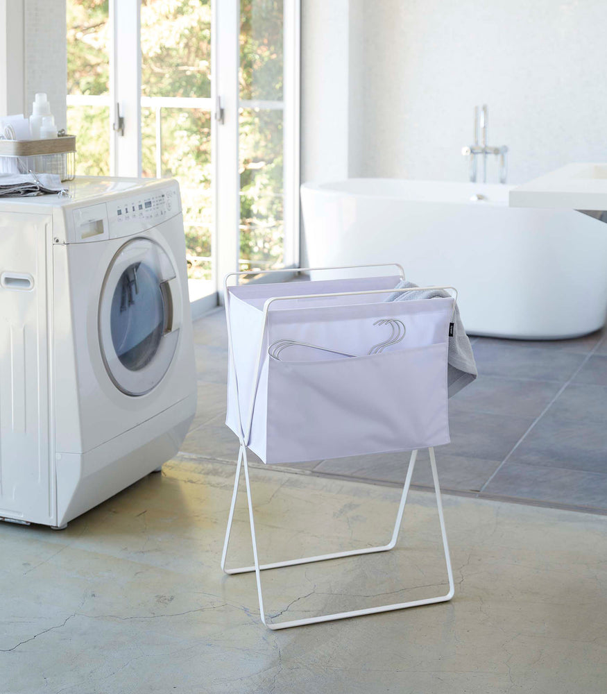 View 14 - A white laundry hamper with white metal legs is angled in front of a washing machine. Wired hangers are poking out of a pocket in the front of the hamper and a towel is seen inside. A washing machine and bathtub are visible in the background.