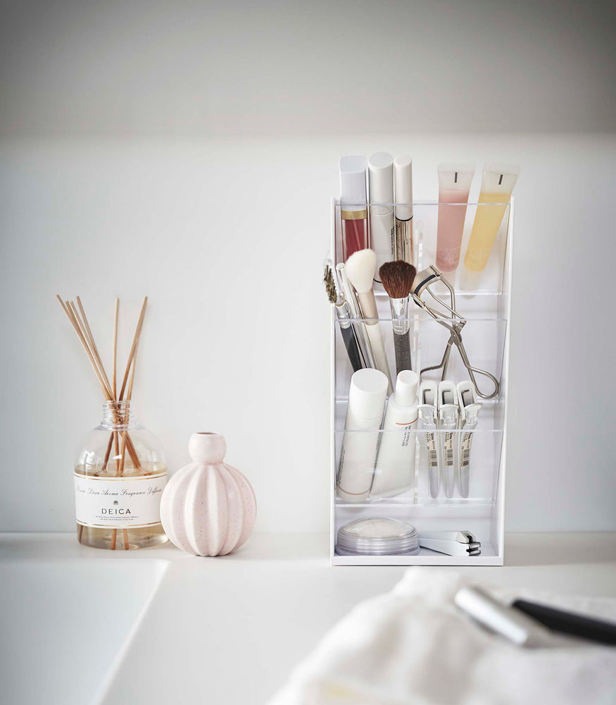 View 3 - A front view of a white rectangular resin cosmetics organizer on a white bathroom counter. It has three deep transparent trays that sit diagonally with adjustable transparent dividers placed in the middle of each tray. The top tray holds lipsticks and glosses
