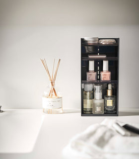 A black resin rectangular cosmetics organizer sits on a white bathroom counter. The organizer has three transparent shelves with upward facing lips to prevent products from falling-out. view 30