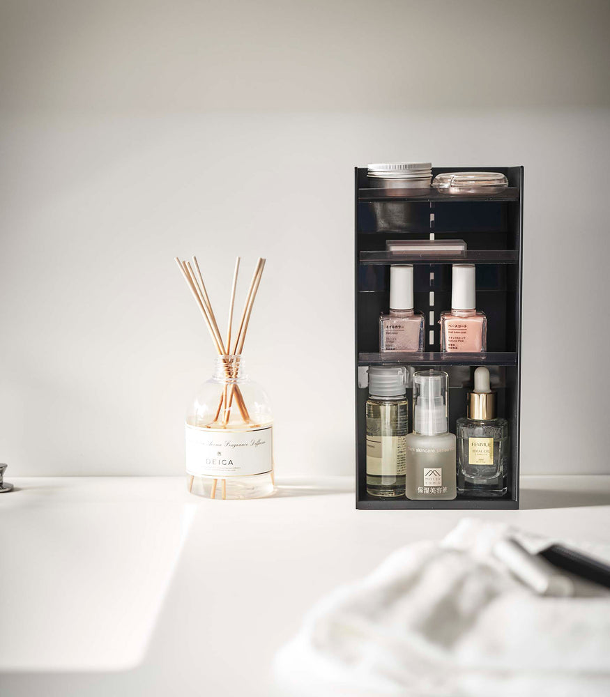 View 30 - A black resin rectangular cosmetics organizer sits on a white bathroom counter. The organizer has three transparent shelves with upward facing lips to prevent products from falling-out.