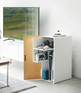 A white video game storage cabinet is placed in a living room next to the TV. The wooden door is open, showcasing organized video games and controllers on two sliding shelves view 3