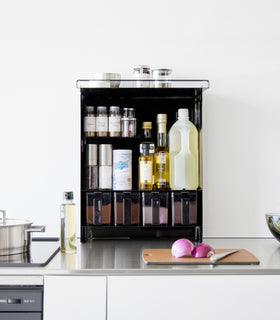 Front view of the Concealable Spice Rack in black by Yamazaki Home with the front panel open, showing various spices and condiments inside. view 15