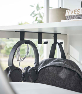 Three matte black metal hooks are secured to the underside of white desk. One hook holds a pair of over-ear headphones and a wrapped charger cord, while another holds a backpack. view 7