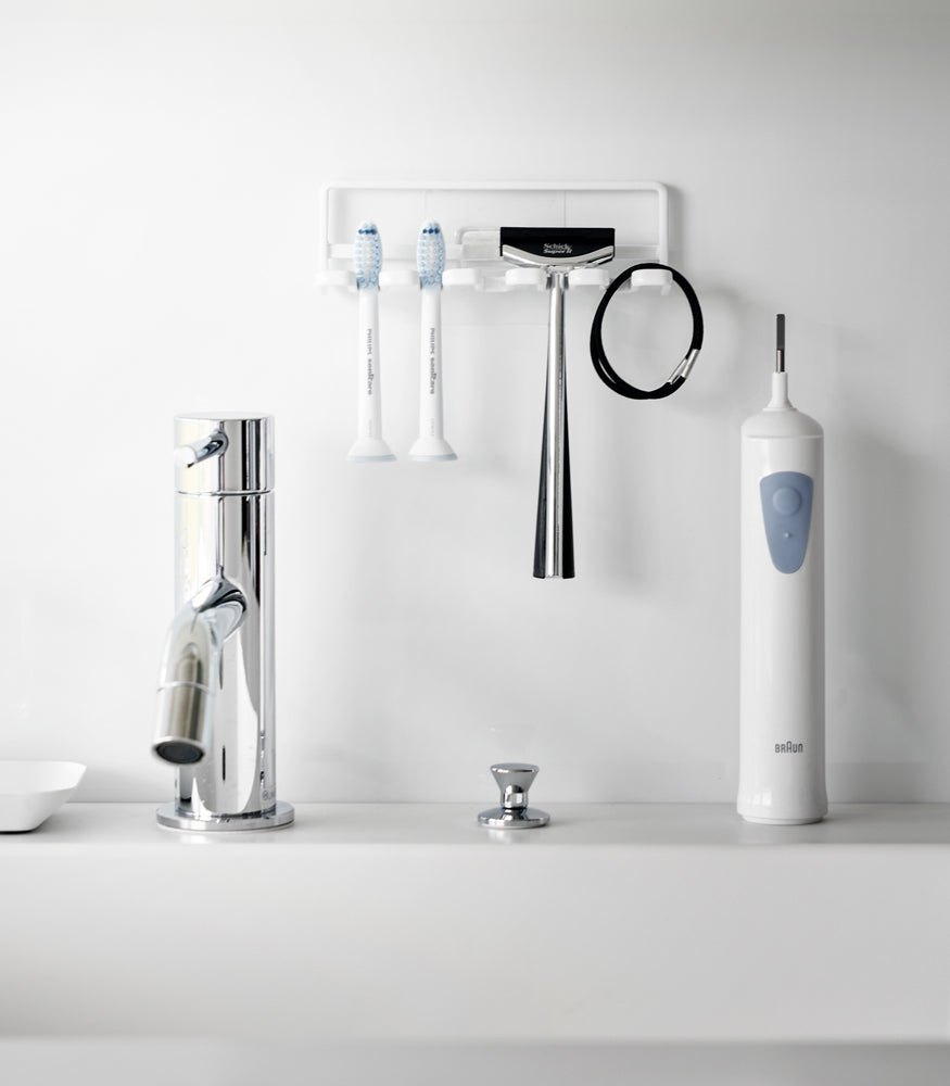 View 4 - Yamazaki Home's white Traceless Adhesive Toothbrush Holder on a bathroom wall, holding two toothbrushes and a razor beside a faucet.