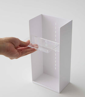 A male hand pulls a transparent tray to adjust the location in a cosmetics organizer. It is a white resin rectangular cosmetics holder with an open face and top. The removable tray acts as a shelf and has an upward facing lip along the edge to prevent products from falling out. view 27