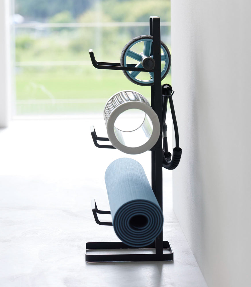 View 16 - Side view of Yamazaki Home Black Fitness Equipment Storage Rack holding a yoga mat and foarm rollers.