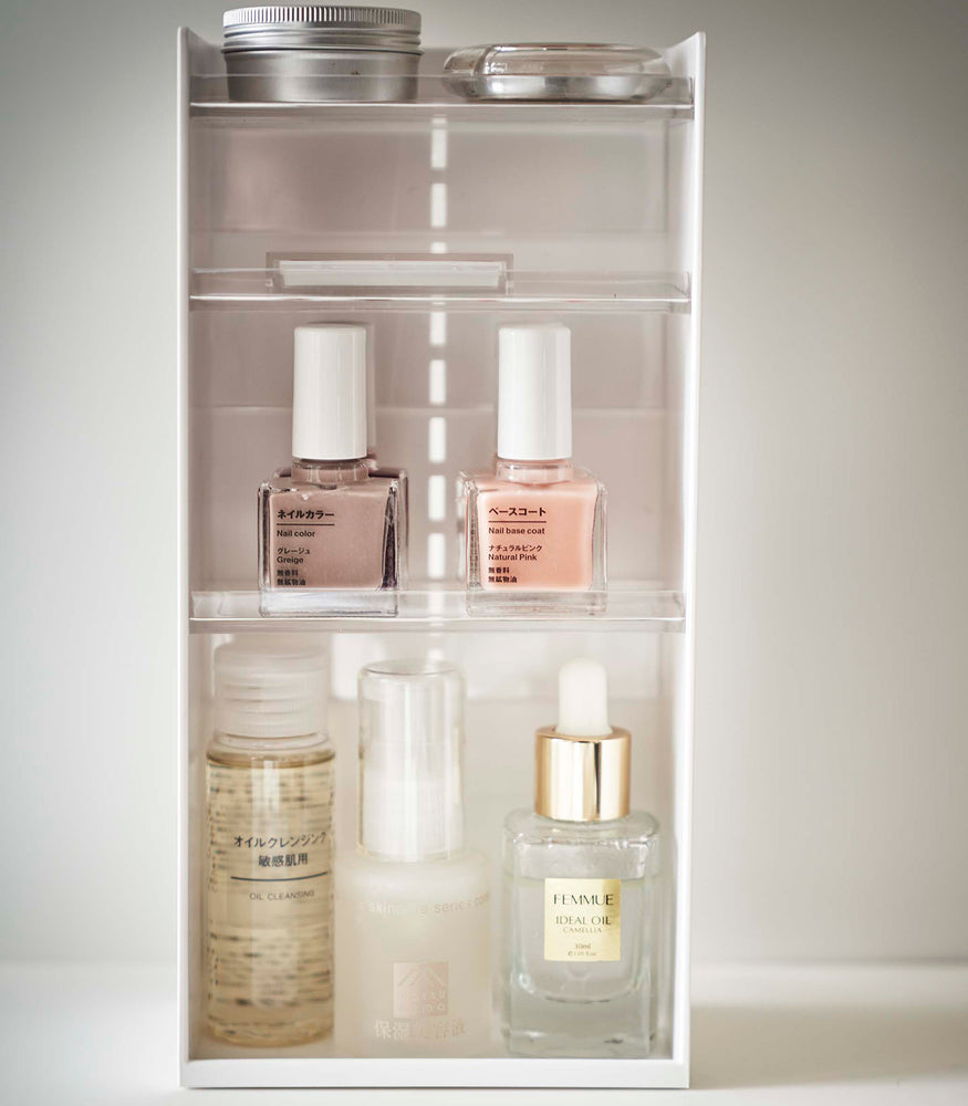 View 7 - On top of a white surface is a white resin rectangular cosmetics organizer with an open face and top. It has three transparent shelves with upward facing lips to prevent products from falling-out.