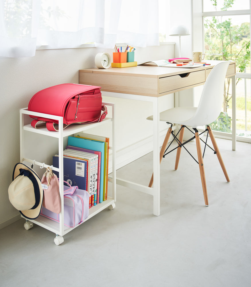 View 5 - Two-level white steel storage cart with casters is shown next to a desk. A backpack is shown on the upper shelf and the lower shelf houses books and a small bag. Three hooks are installed on one side, holding a hat and a small bag. One side of the cart has a wall to hide storage when flipped.