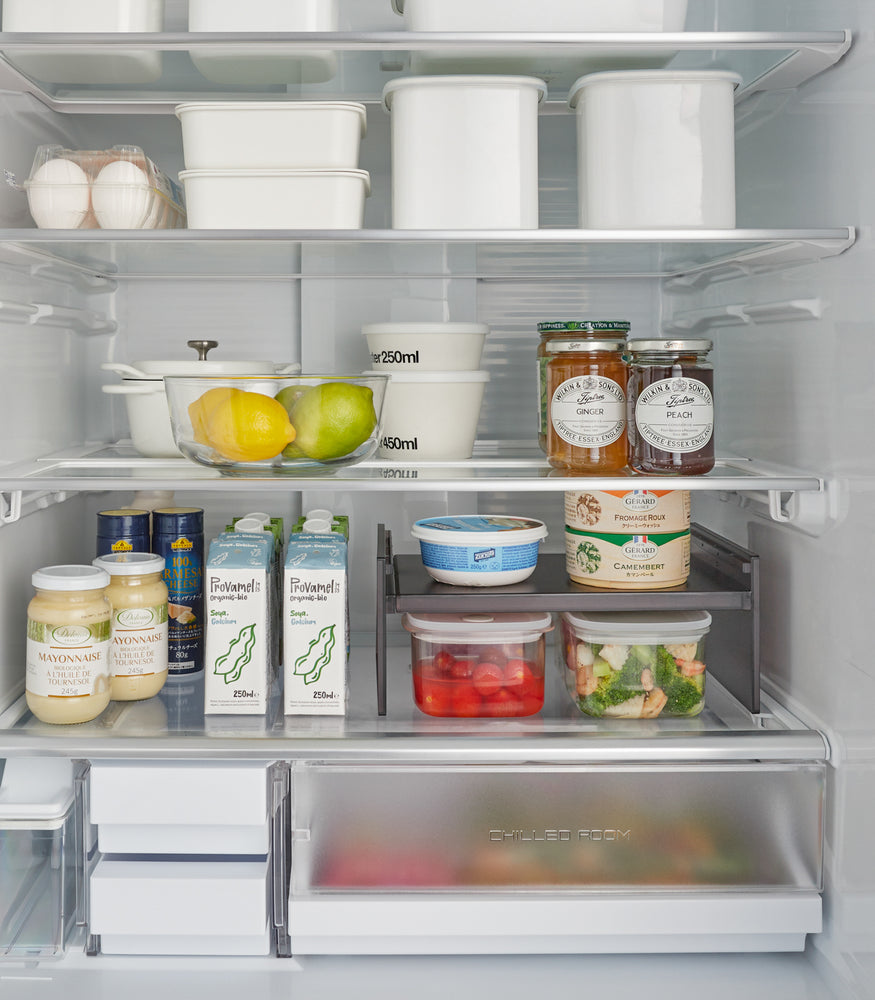 View 13 - Black Expandable Refrigerator Riser Shelf by Yamazaki Home featured in a refrigerator, helping to organize and separate various food products.