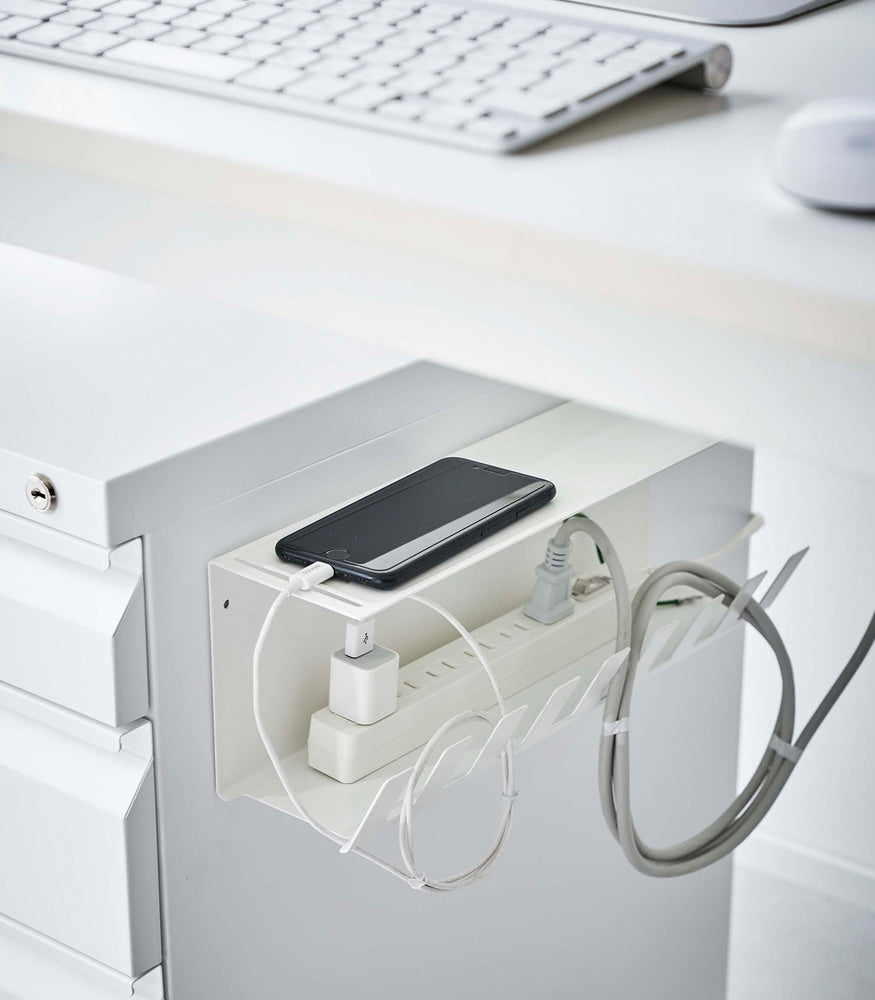 View 8 - The Magnetic Under-Desk Cable Organizer in white by Yamazaki Home mounted on the side of a file cabinet holding a power strip and phone.