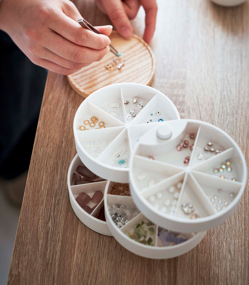 View 13 - A bird’s eye view of a white four-tier jewelry organizer. The organizers tiers are swiveled in a way so that the inside contents are visible, each tier is divided into seven individual compartments in a pie-shaped fashion. Each individual compartment of the tiers holds various beads and jewelry-making parts. Behind the organizer is the light-colored wooden lid with a person using tweezers to pick up a bead. Around the lid is a raised lip.