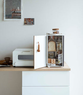 A vertical white metal breadbox is seen on a wooden kitchen counter next to a white microwave oven. The breadbox’s door is swung open to the left and bread, other grains, and a bottle of wine are seen inside the box. A magnetic stop is seen opposite the open door. On the open door is a white hook holding brown plastic measurement spoons. On top of the box is a folded towel and plastic container of cookies. view 11