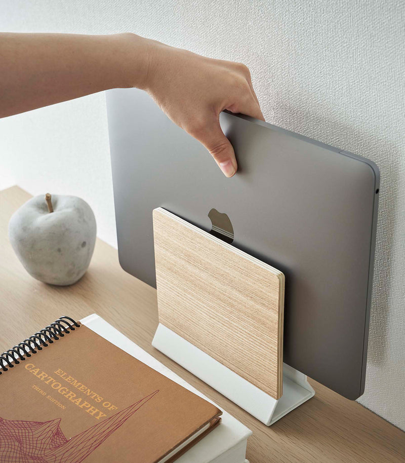 A hand lifts a closed laptop from a narrow wooden stand with a white metal base positioned on top of a desk.