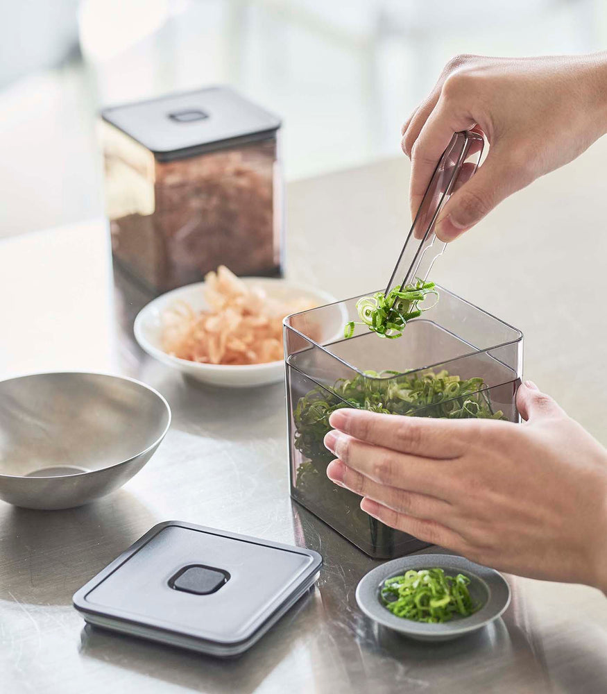 View 11 - Person using tongs to grab scallions out of black Vacuum-Sealing Food Container on kitchen countertop by Yamazaki Home.