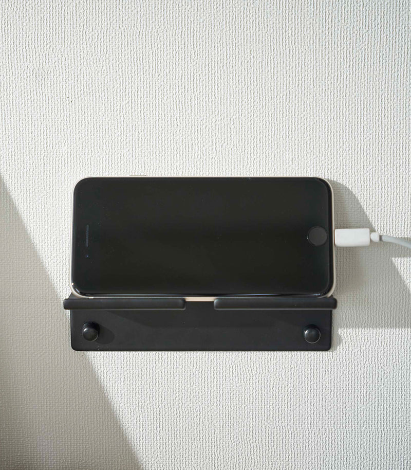 An iPhone with a charger cord plugged in is mounted on a wall. The screen is facing outward and the phone is held up on its side.
