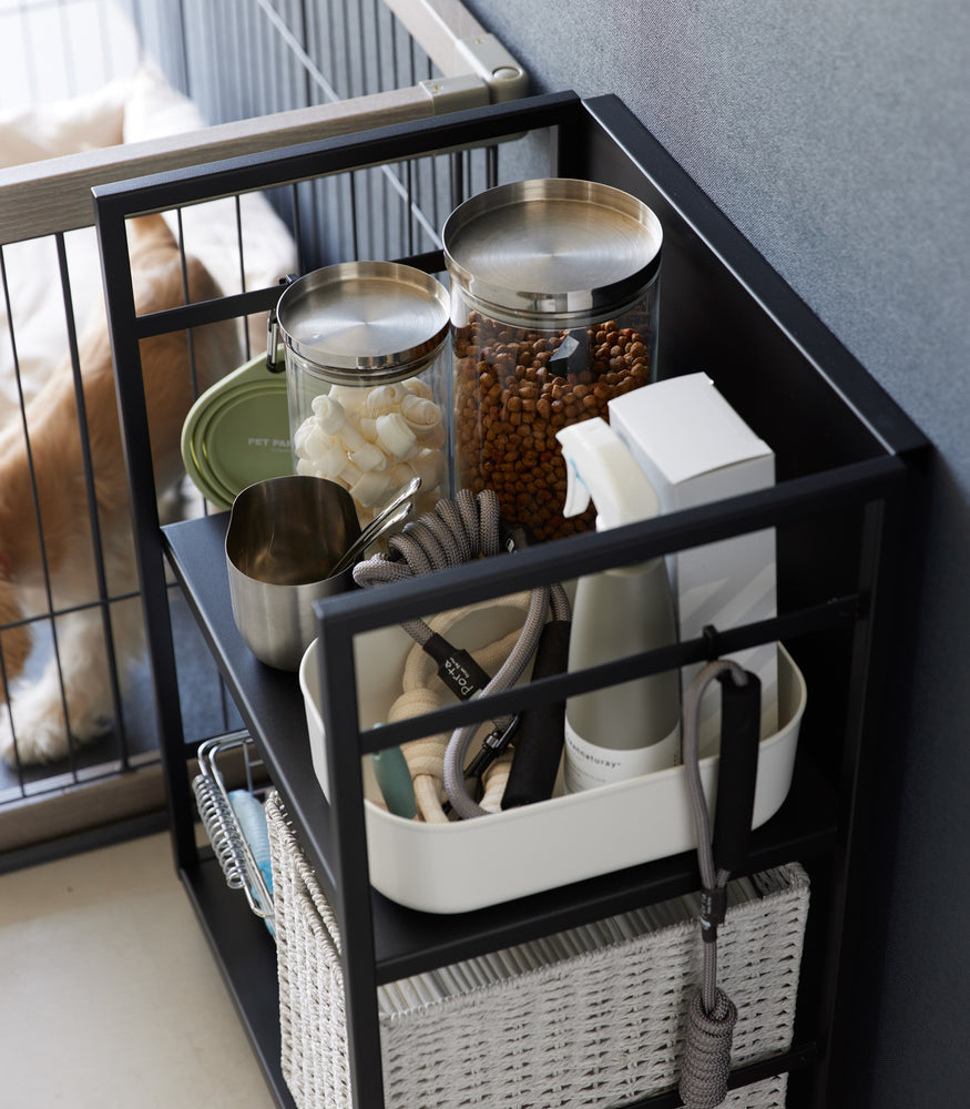 View 16 - Two-level black steel storage cart with casters is shown next to a dog crate. Cart houses various dog supplies and treat storage on the upper shelf, bottom shelf holds two storage bins. Hooks are installed on both sides, one holds a collapsible water bowl, the other a leash. Solid back wall of the cart demonstrates that the items can be hidden when flipped.