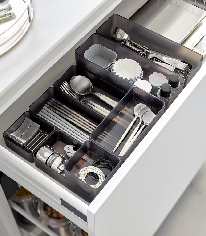 View 64 - Four different sizes of stackable clear organizers in black —small, medium, large, and deep—are neatly placed in a kitchen drawer. They effectively organize various items such as cutlery, bottle openers, and small trays, showcasing their versatility and stackability