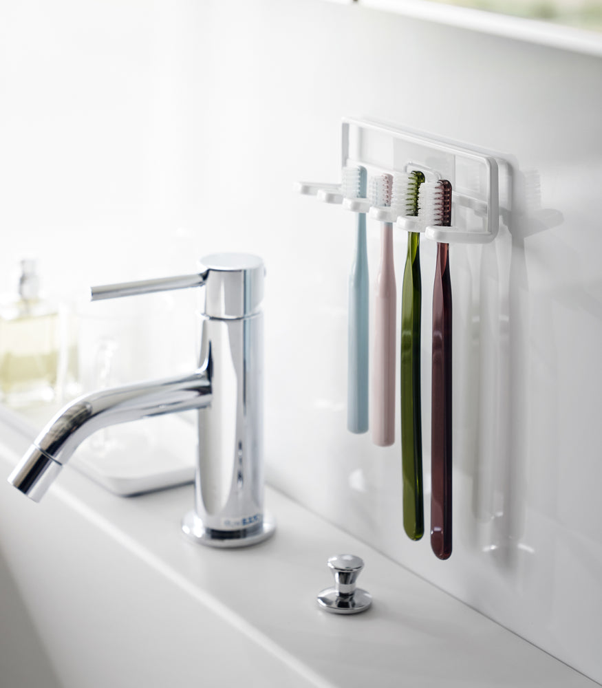 View 2 - Yamazaki Home's Traceless Adhesive Toothbrush Holder, white, mounted on a bathroom wall with four colorful toothbrushes and a chrome faucet.