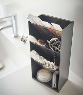 An upper view of a black rectangular resin cosmetics organizer on a white bathroom counter. It has three deep black transparent trays that lean forward diagonally with matching dividers placed in the middle of each tray. The middle tray is in focus and holds a set of cosmetic brushes and an eyelash curler. In the background is an out-of-focus sink faucet and basin. view 16