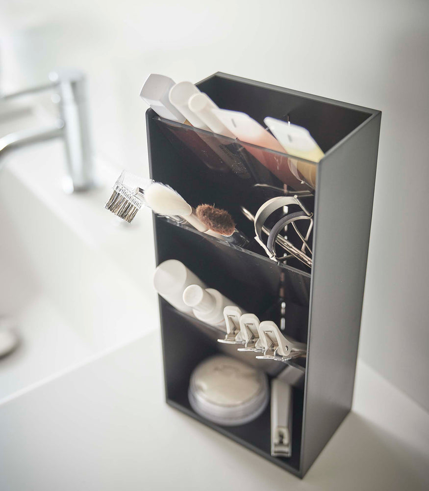 View 15 - An upper view of a black rectangular resin cosmetics organizer on a white bathroom counter. It has three deep black transparent trays that lean forward diagonally with matching dividers placed in the middle of each tray. The middle tray is in focus and holds a set of cosmetic brushes and an eyelash curler. In the background is an out-of-focus sink faucet and basin.