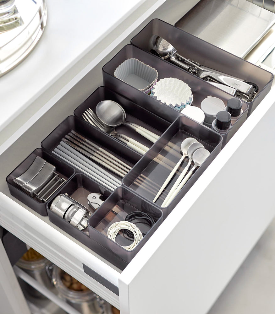 View 60 - Four different sizes of stackable clear organizers in black—small, medium, large, and deep—are neatly placed in a kitchen drawer. They effectively organize various items such as cutlery, bottle openers, and small trays, showcasing their versatility and stackability