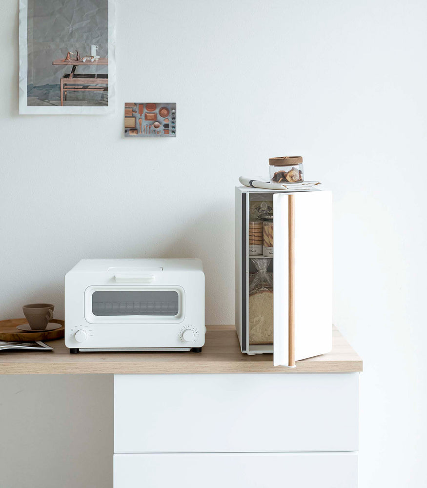 View 9 - A vertical white metal breadbox, with a vertical wooden handle, is seen on a white kitchen counter next to a white microwave oven and a wooden plate with a brown coffee cup and matching saucer on it.