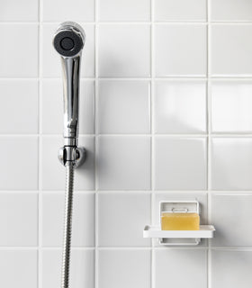 Yamazaki Home's white Traceless Adhesive Soap Tray in a shower, holding a bar of soap next to a silver shower hose on tiled wall. view 7