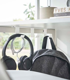 Three matte white metal hooks are secured to the underside of white desk. One hook holds a pair of over-ear headphones and a wrapped charger cord, while another holds a backpack. view 2