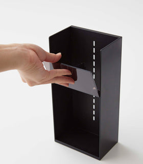 A male hand adjusts a diagonally sloped black transparent tray’s location on a cosmetics organizer. It is a black resin rectangular cosmetics holder with an open face and top. Small evenly distributed rectangular cut-outs go down the middle of the organizer, allowing the trays locations to be adjusted. The bottom of the organizer has a slight upward pointing lip. view 9