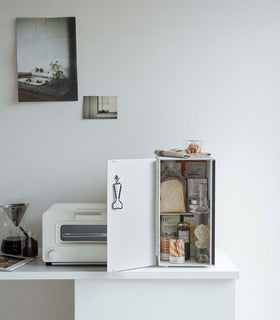A vertical white metal breadbox is seen on a white kitchen counter next to a white microwave oven and a glass drip coffee pot with a dark liquid inside. view 17